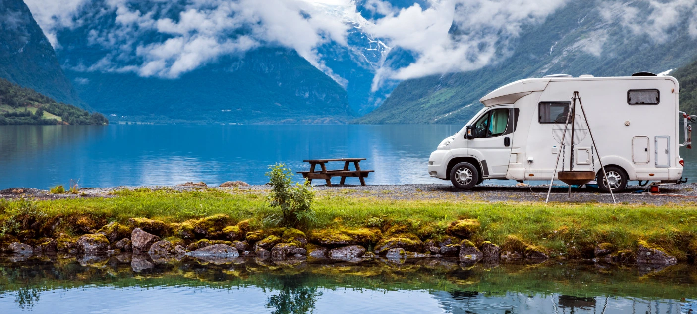 Wohnmobil am See mit Bergen und Wolken im Hintergrund, Picknickbank und Feuerstelle daneben.